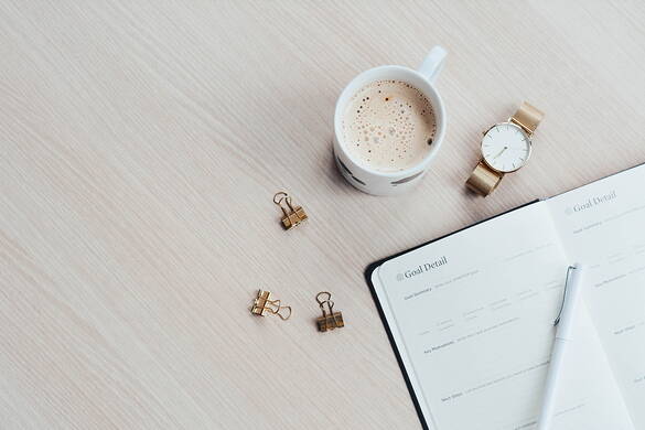 Eine Tasse mit Milchkaffee steht auf dem Tisch neben einer Uhr, drei Büroklammern und einem Journal.