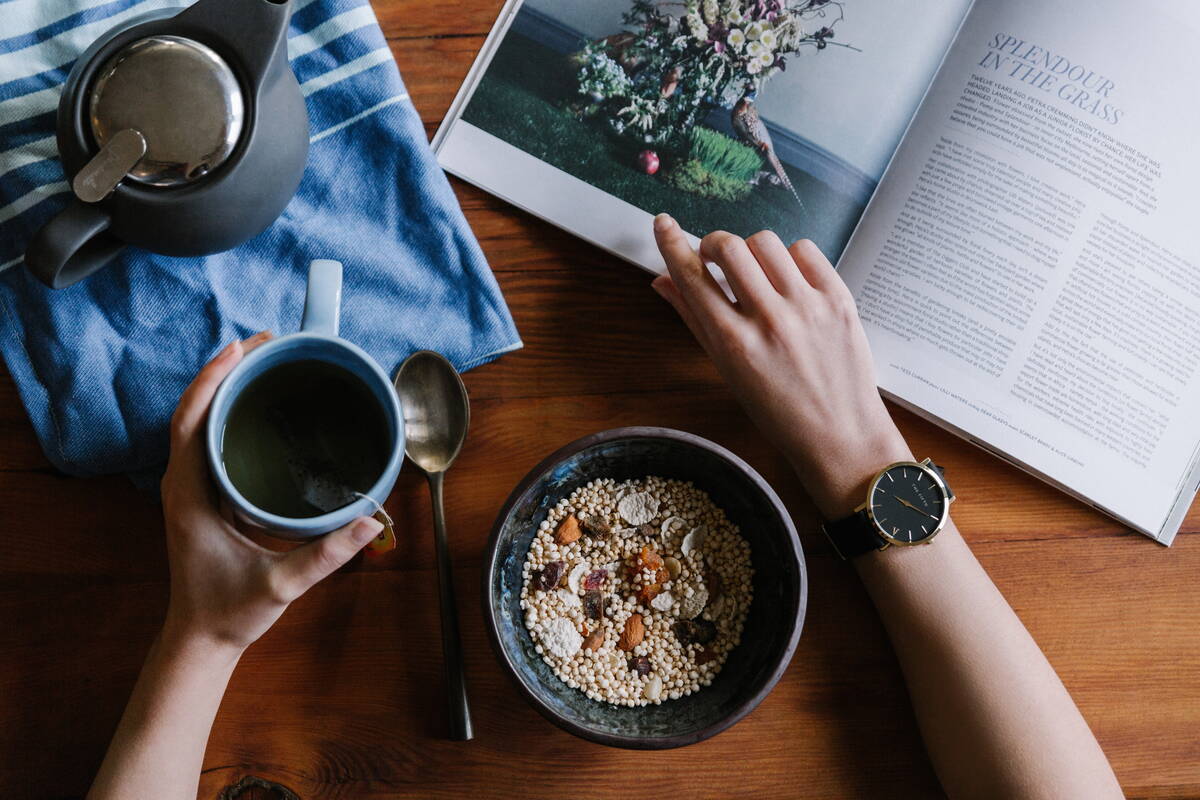 daughters-of-time_bulletjournal_timemanagement Eine weibliche Person sitzt an einem Holztisch, zu sehen sind allerdings nur ihre beiden Unterarme und ihre Hände. In der linken Hand hält sie eine Tasse Tee. Daneben liegt ein Löffel und davor ein blaues Tuch mit weissen Streifen und darauf eine Teekanne. Rechts vom Löffel steht eine Müslischale und die rechte Hand liegt ruhig noch weiter rechts daneben. Am Handgelenk trägt die Frau eine schwarze Uhr mit goldenen Ziffern und die Hand ruht auf einem Magazin mit einem Bild von Blumen.