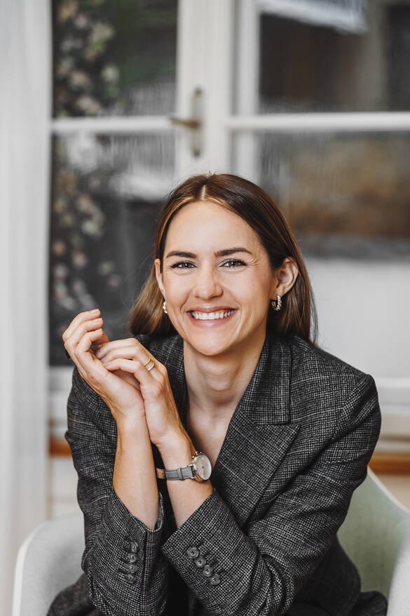 Chantal Muri sitzt auf einem Stuhl vor einem Fenster. Das Fenster ist verschwommen, der Fokus liegt auf ihr. Sie lächelt und hat ihre Ellenbogen auf ihren Beinen abgestützt. Sie trägt eine graue Armbanduhr, einen karrierten Blazer und zwei silberne Creolen. Ihre Haare trägt sie offen und hinter ihren Ohren. 