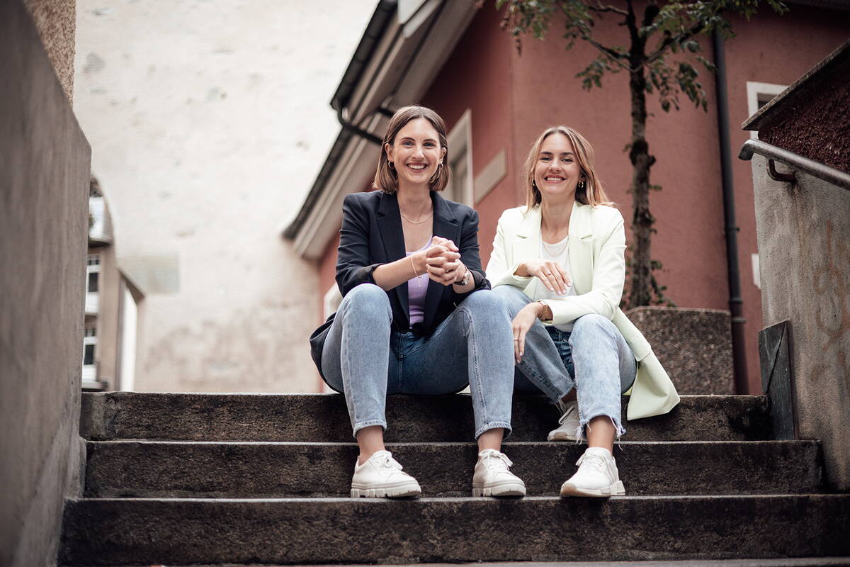 Zwei jugen Frauen sitzen auf einer Treppe und lachen in die Kamera.