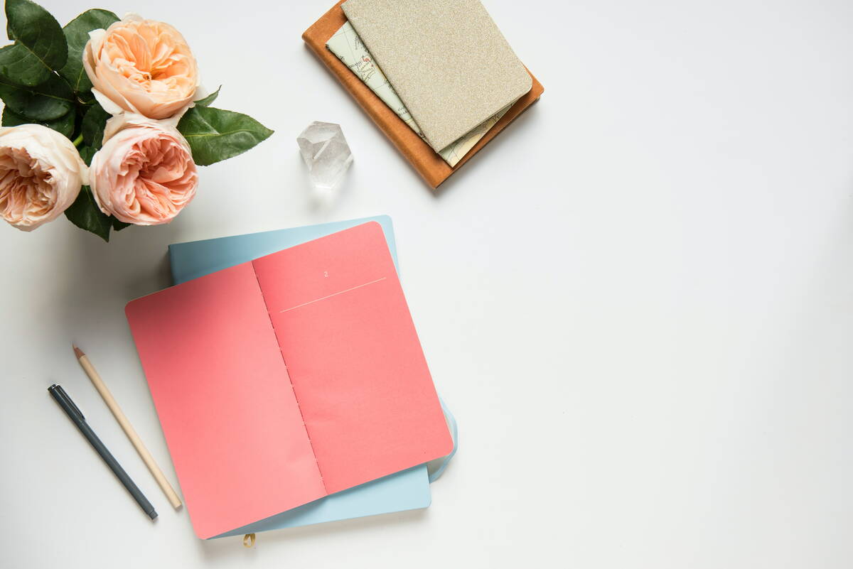 daughters-of-time_bulletjournal_timemanagement Auf weissem Grund stehen verschiedene Objekte, die von oben fotografiert wurden. In der Ecke links des Bildes stehen drei Rosen inklusvie grünen Blättern. Daneben rechts liegt ein Bergkristall. Nochmal etwas weiter rechts liegen drei Notizbücher aufeinander, so dass alle zu sehen sind. Vor dem Bergkristall, weiter unten im Bild, liegen nochmal zwei Notizbücher. Eines liegt geöffnet da. Zwei Stifte liegen links davon.