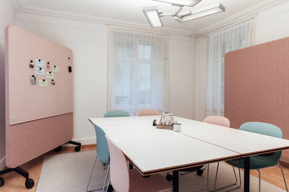 Unser Seminarraum mit drei Tischen, sechs Stühlen und zwei Glassboards. Der Raum ist hell und freundlich eingerichtet in sanften rosaroten und türkis Tönen.