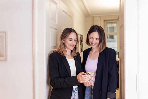 Chantal Muri und Fabienne Muri stehen in ihrer Coachingpraxis im Türrahmen und schauen gemeinsam auf ein Handy. 