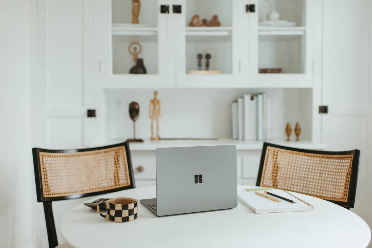 Ein Laptop steht in der mitte eines weissen rundne Tisches. Daneben liegen ein Notizheft und zwei Stifte, ein Taschenrechner und eine Tasse. Das Bild ist sehr symetrisch und in ruhigen braun, schwarz und weiss Tönen gehalten. 