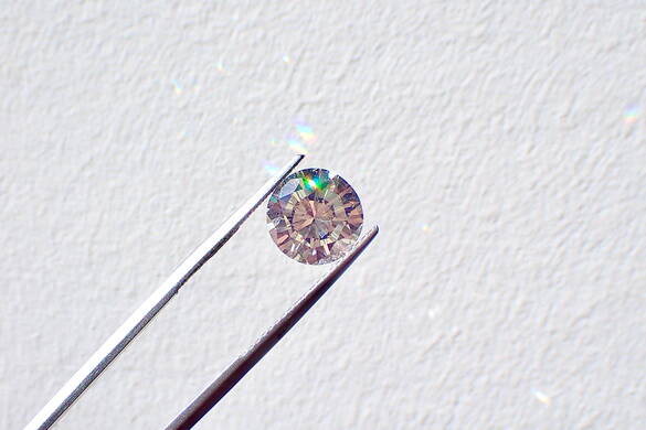 brillieren_brilliant_sein.jpg Eine feine Zange hälte einen Diamant vor einer weissen Wand.