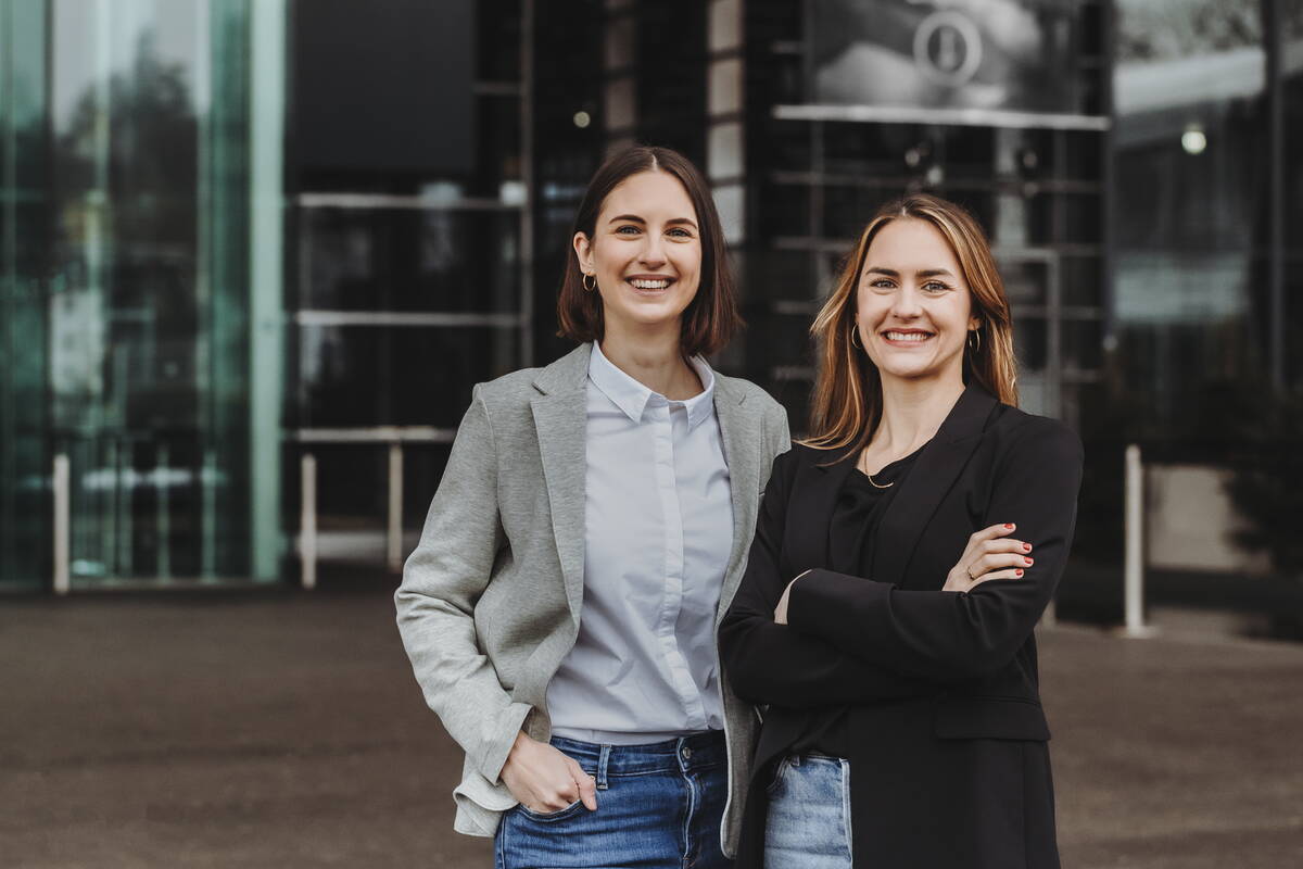 Fabienne Muri und Chantal Muri stehen nebeneinander vor einem modernen Glashaus und lachen direkt in die Kamera. Chantal hat ihre Hände vor der Brust verschränkt, Fabienne hat eine Hand in ihrer Hosentasche verstaut. Chantal trägt Jeans, einen schwarzen Blazer, schwarzes T-shirt, roten Nagellack, goldene Creolen, eine Goldkette und einen goldenen Ring. Fabienne trägt eine Jeans, eine weisse Bluse und einen grauen Blazer. 