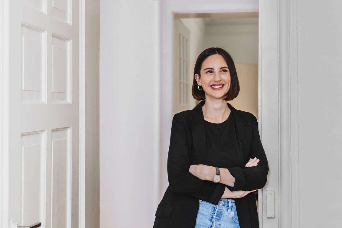 Fabienne steht in einem alten Haus in einem weissen Türrahmen und lächelt an der Kamera vorbei. Sie trägt eine Jeans, ein schwarzes T-shirt, einen schwarzen Blazer und eine braune Uhr. Ihr Haare sind braun und kinnlang. Die Hände hat sie locker vor der Brust verschränkt. 